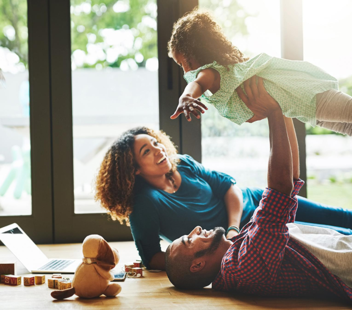 Imagem de uma família, composta por: um casal e uma criança deitados no chão brincando
